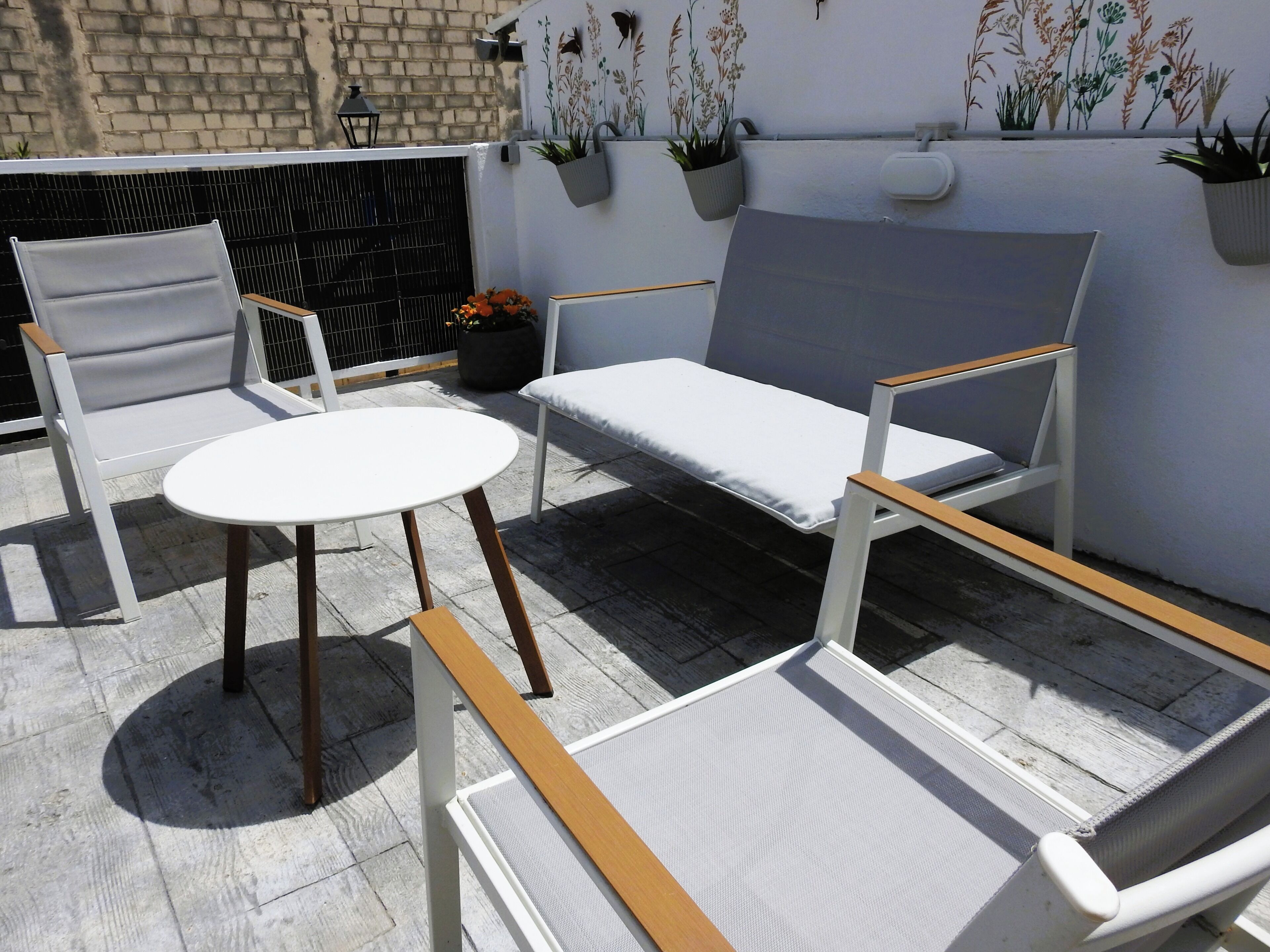 Un patio con una mesa blanca y dos sillas, un banco y plantas en macetas frente a una pared con decoración floral.