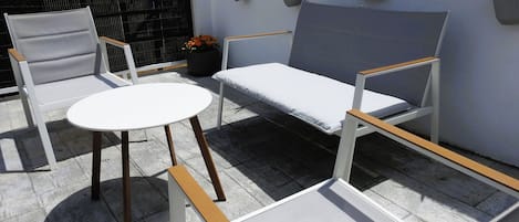 Un pati amb una taula blanca i dues cadires, una banca i plantes en testos davant d'una paret amb decoració floral.