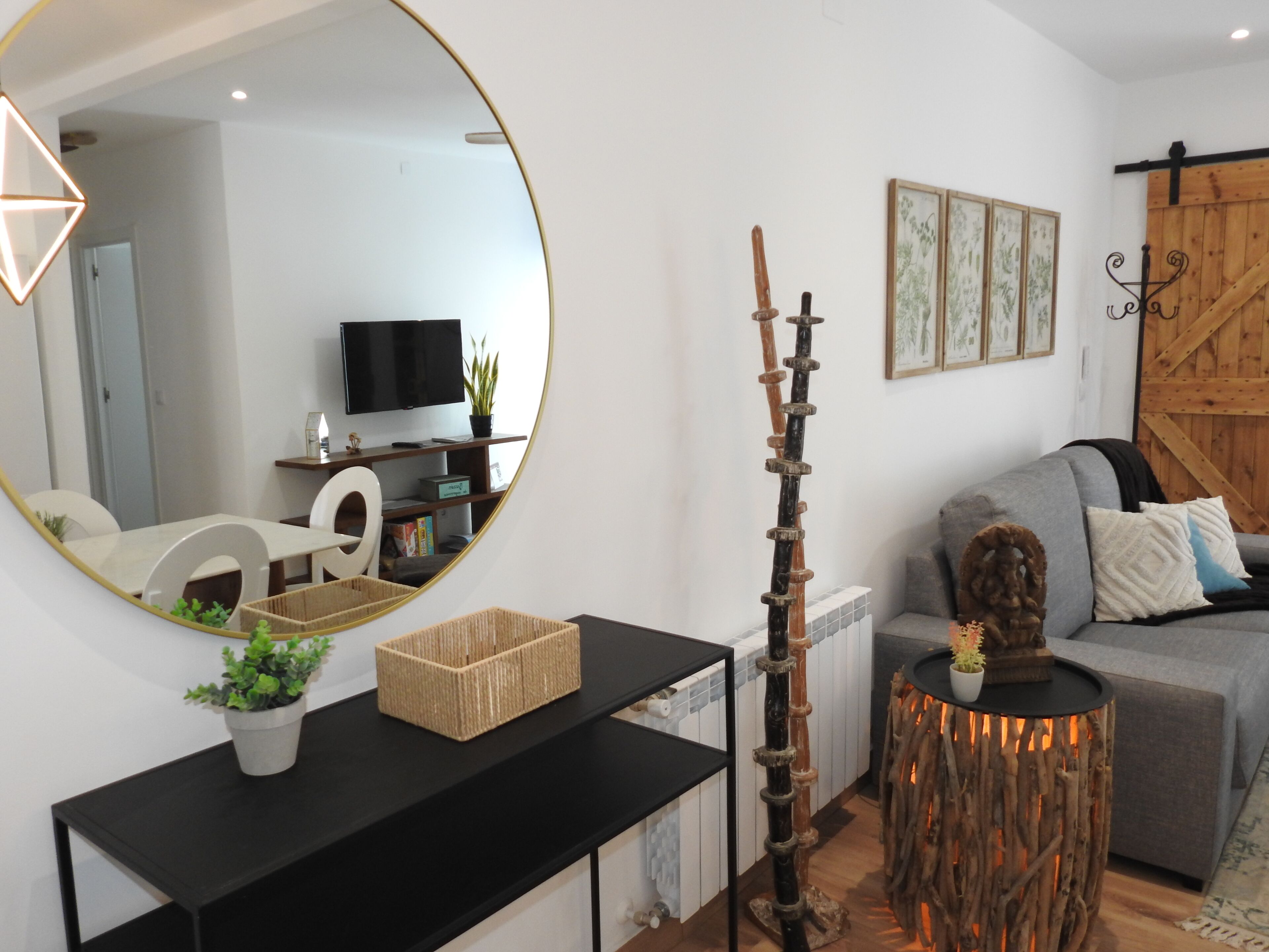 Una sala moderna con un espejo redondo, un adorno de madera, un sofá gris y un televisor sobre un mueble.