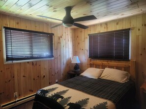 A room with wooden walls, a bed with a plaid bedspread, two windows with blinds, a ceiling fan, and a nightstand with a lamp.