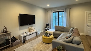 En moderne stue med et fladskærms-tv, en sofa, en guldfarvet skammel og et stort vindue med gardiner.