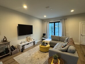 Ein modernes Wohnzimmer mit Fernseher, Sofa, einem gelben Hocker und einem großen Fenster mit Vorhängen.