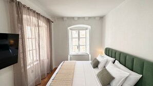 Ett sovrum med en säng, en tv, ett fönster med gardiner och en grön, klädd sänggavel.