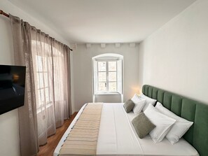 Quarto com cama, televisão, janela com cortinas e cabeceira estofada verde.