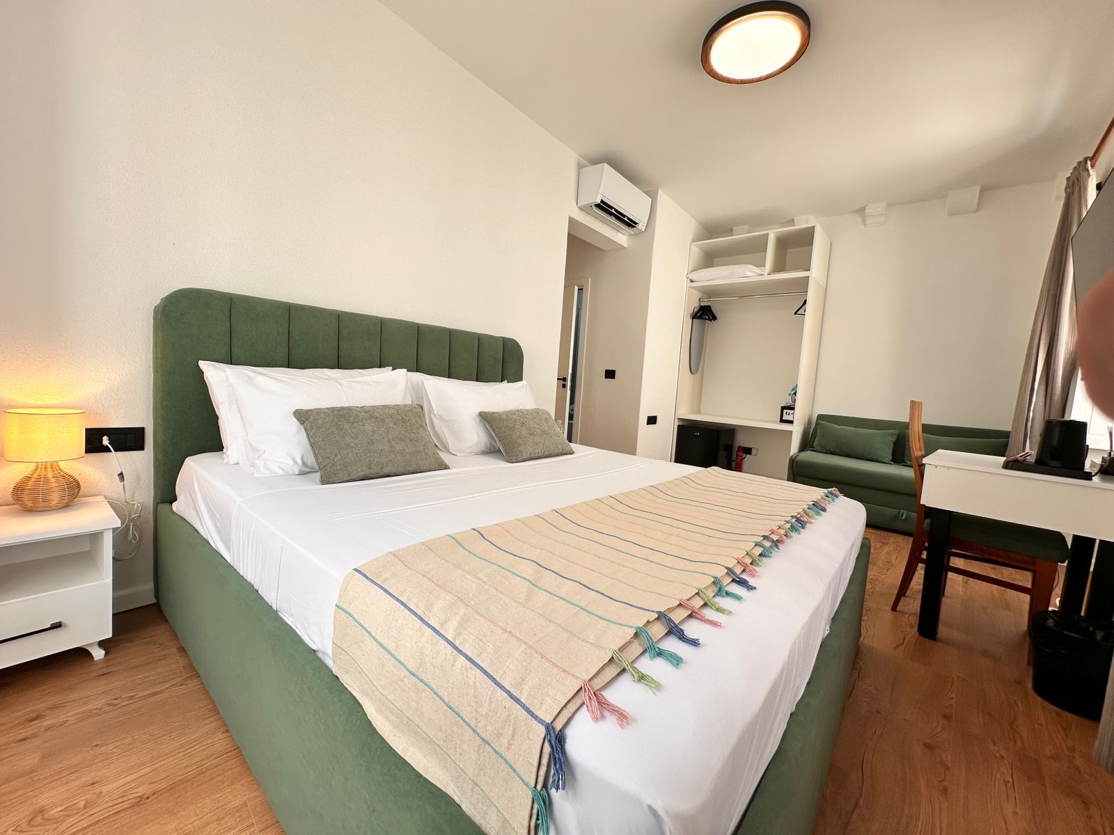 Quarto de hotel com cama, mesa de cabeceira, abajur, guarda-roupa e secretária com cadeira.