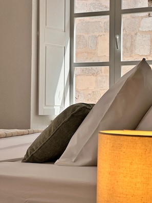 Una cama con almohadas y una lámpara en una habitación con ventana y pared de piedra.
