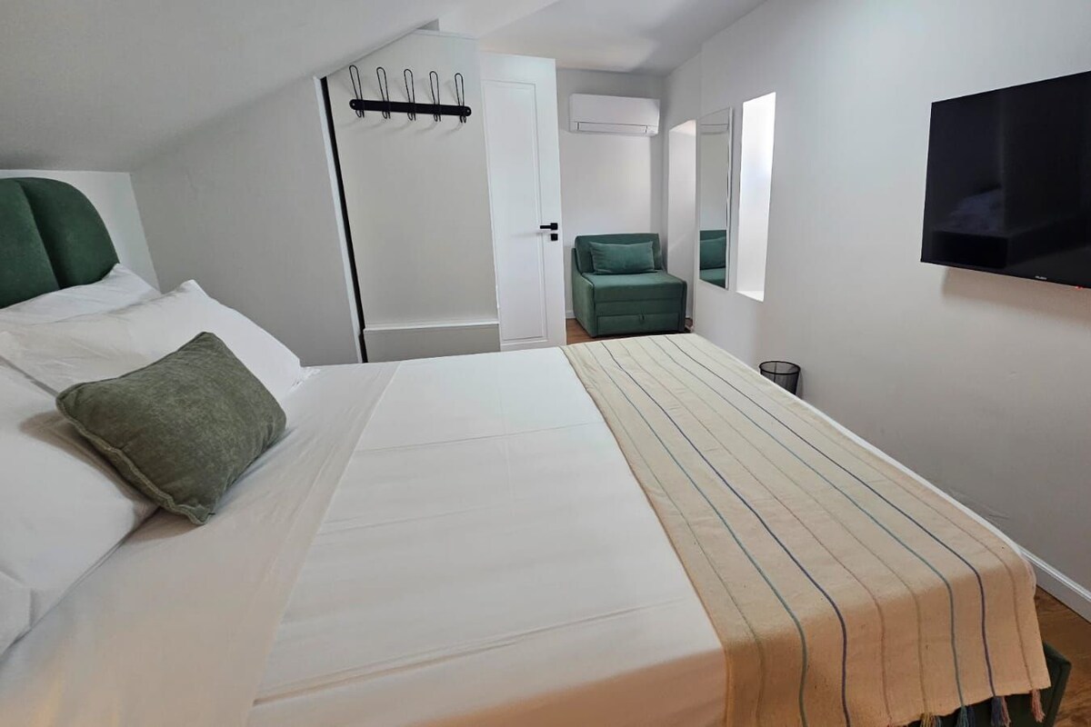 Quarto de hotel com cama, uma cadeira verde, televisão e porta com cabideiro.