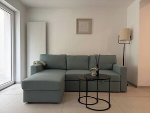 Uma sala de estar moderna com um sofá seccional cinza, uma mesa redonda pequena com um vaso e velas, um abajur e um quadro na parede.