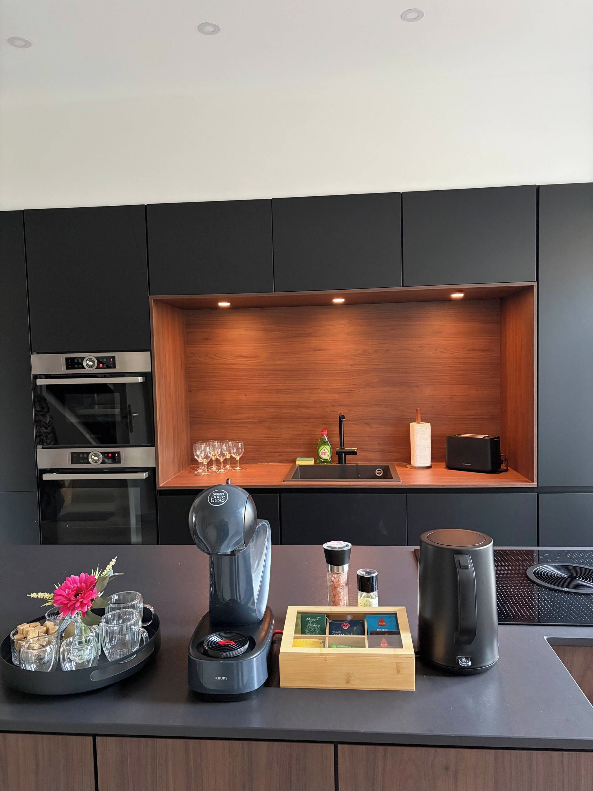 A modern kitchen with built-in appliances, a wooden shelf, and a coffee station.