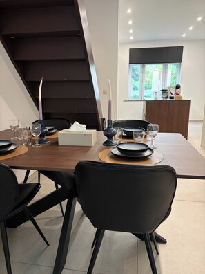 Uma sala de jantar moderna com mesa de madeira posta para quatro, cadeiras pretas e uma escada ao fundo.