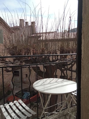 Un balcó amb taula blanca i cadires metàl·liques, amb vista a un pati amb arbres sense fulles i un cotxe estacionat.