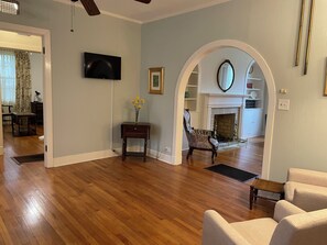 A spacious living room with hardwood flooring, a television mounted on the wall, a fireplace, and a comfortable seating area. - Walk to MSU, Downtown & Cotton District  Historic Home, Sleeps 6 (Starkville)
