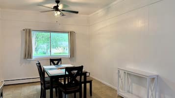 Un coin repas comprenant une table, des chaises, un ventilateur de plafond, une fenêtre avec des rideaux et un radiateur blanc.