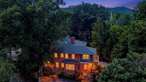 Une maison à deux étages, entourée d’arbres denses, avec une cheminée bien visible et une entrée lumineuse.