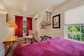 Un dormitorio con cama, una silla, una mesa, una ventana con cortinas, estante colgante y un espejo instalado en la pared.