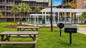 Un'area verdeggiante con tavoli da picnic e un barbecue, con una piscina e degli edifici sullo sfondo.