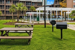 Un área con césped, mesas de picnic y una parrilla, con una piscina y edificios al fondo.
