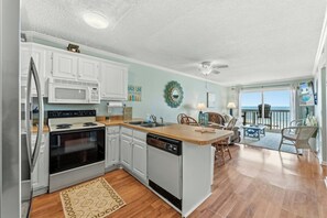 A compact kitchen with white cabinets, a microwave, an oven, and a dishwasher. There's a wooden island with a sink, and a dining area with chairs and a view of the ocean through large windows. - Ocean Garden Villas B3- Oceanfront 2 Bedroom (North Myrtle Beach)