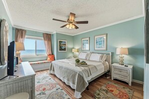 Ein Schlafzimmer mit einem Bett, einem Deckenventilator, einem Fenster mit Vorhängen, einem Nachttisch mit Lampe und einem an der Wand befestigten Fernseher.