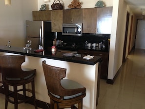 A kitchen with a central island, two bar stools, stainless steel appliances, and wooden cabinets. - 3-bedroom Ocean Front Villa, Dawn Beach Club at JW Marriott. Fixed 6403 (oyster pond)