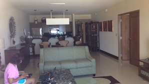 Sala de estar com sofá, mesa de centro e uma pessoa usando um laptop. Cozinha visível ao fundo.