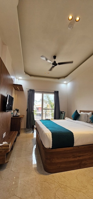 A hotel room with a bed, a desk, a ceiling fan, and a window with curtains. - The Sky Comfort Braj dham (Nathdwara)