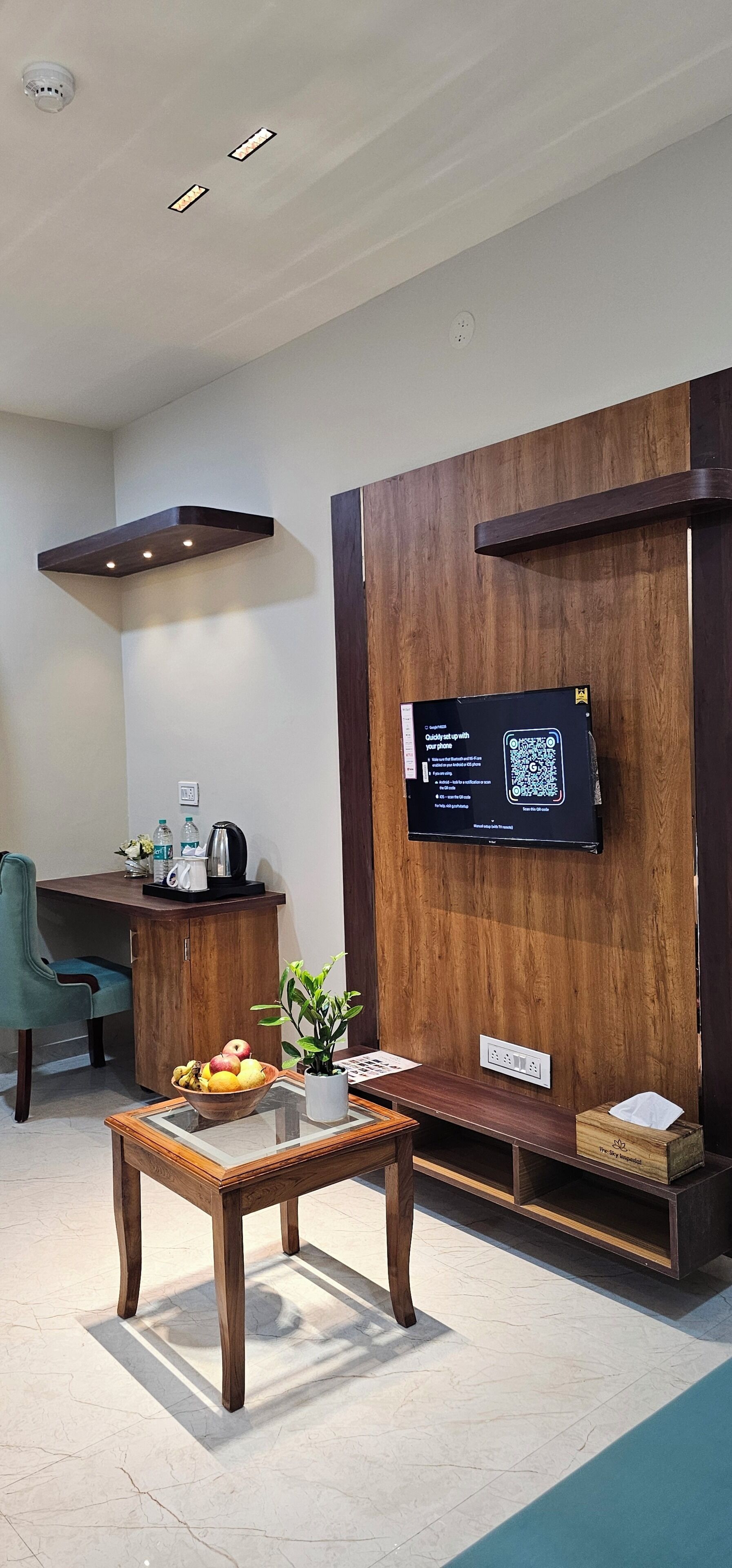 Una habitación moderna con un televisor de pantalla plana montado en una pared de madera, una mesa de madera con un cuenco de fruta, una silla y una pequeña planta en maceta.