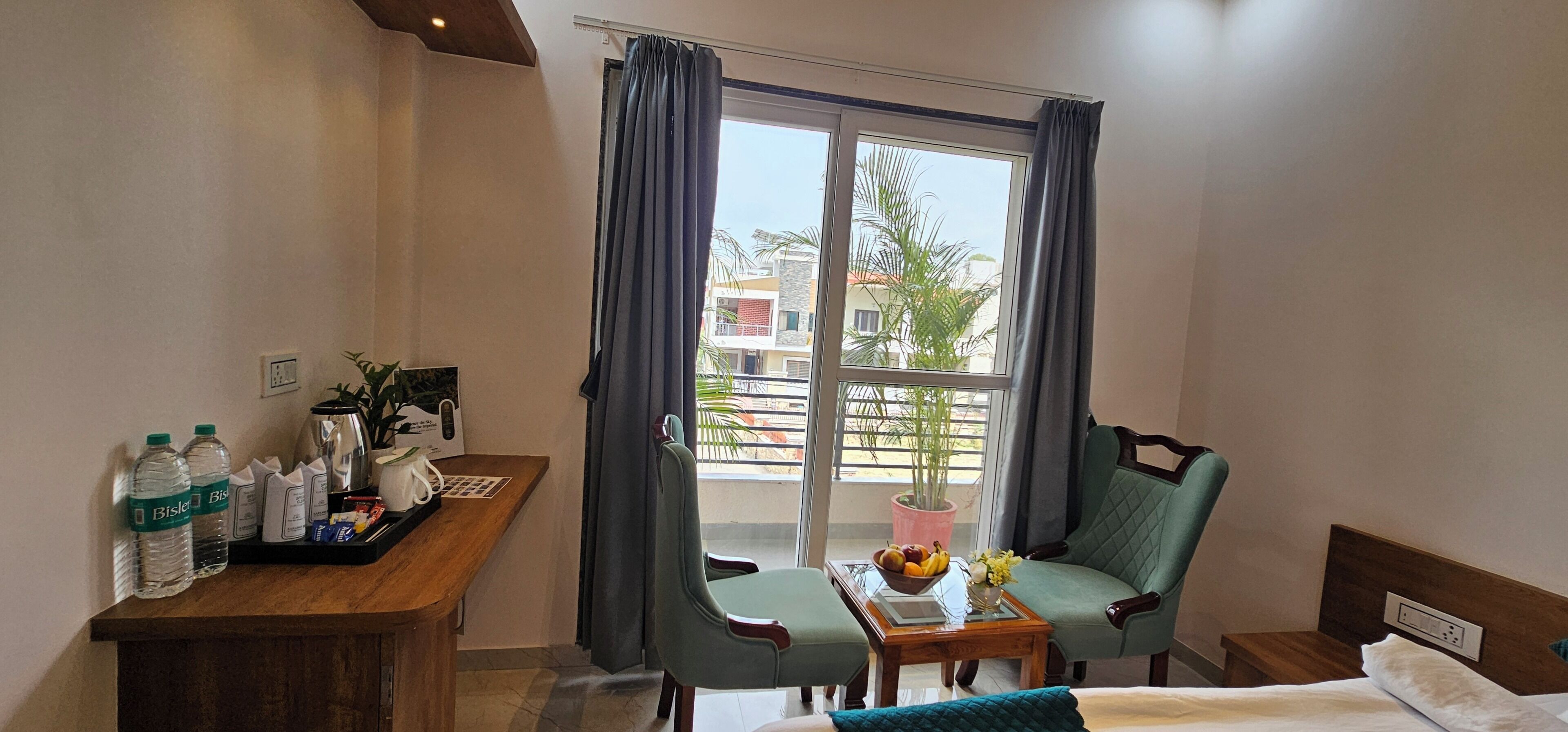 A hotel room with a bed, a desk with toiletries, a chair, a small table with fruit, and a view of the outside.