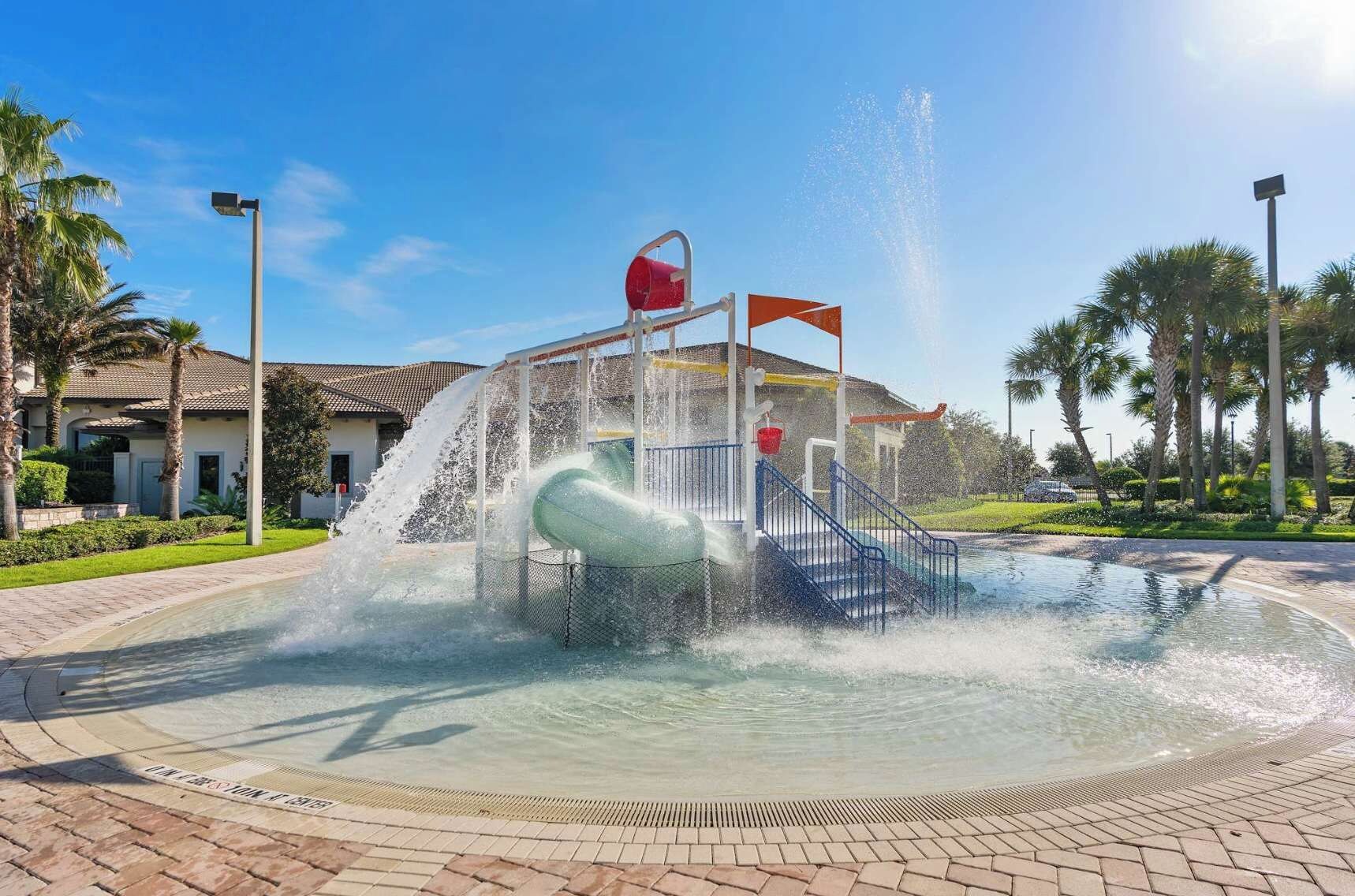 A water play area with a slide and water jets, surrounded by a paved walkway and palm trees.