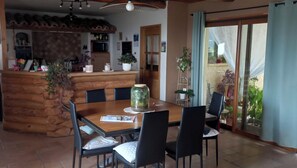 Un coin repas avec une table et des chaises en bois, une cuisine à l’arrière-plan et une porte-fenêtre coulissante donnant sur un espace extérieur agrémenté de plantes.