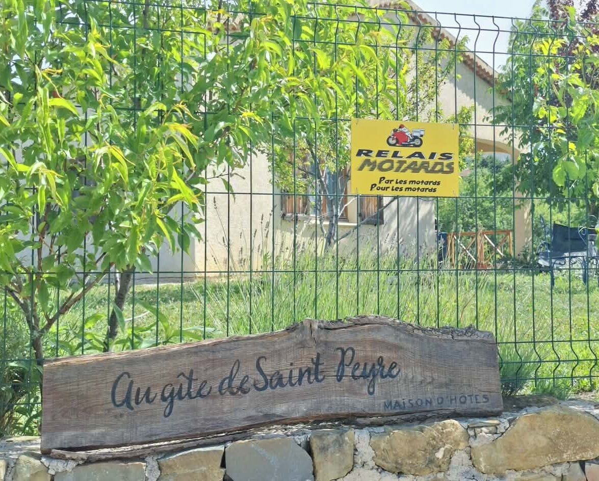 A wooden sign with 'Avenue de Saint Pierre' and 'Mansion Hotel' text, a yellow sign with a motorcycle and text, and a metal fence with greenery behind.