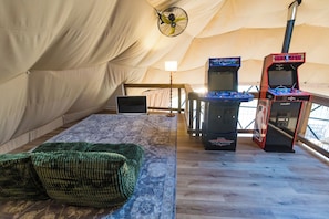 Een tent met een laptop op een tafel, twee speelautomaten, een ventilator en een groene geweven stoel.