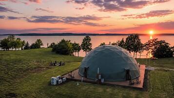 Une structure en forme de dôme géodésique, dotée d’un toit plat, entourée d’un champ verdoyant, près d’une étendue d’eau, avec un coucher de soleil en arrière-plan.