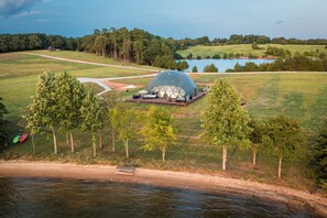 A geodesic dome structure with a patio area, surrounded by a natural landscape with trees and a body of water. - Lake Monticello Geodome-MiniGolf-Hot Tub-Games-Playground-Stargazing-Lake Murray (Jenkinsville)