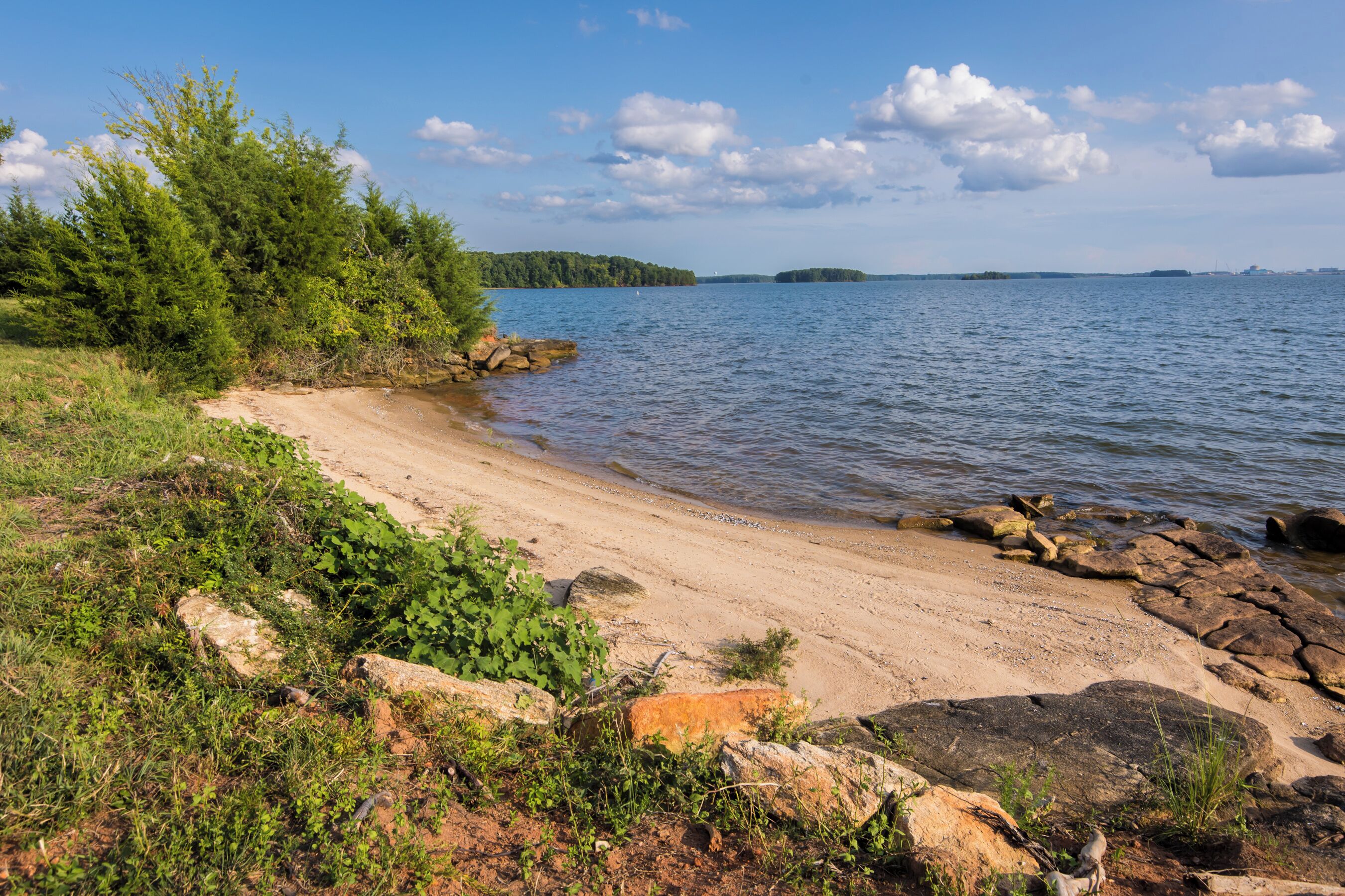 A sandy beach with rocks and greenery, a calm lake, and distant islands.