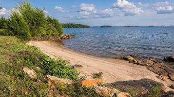 A sandy beach with rocks and greenery, a calm lake, and distant islands.