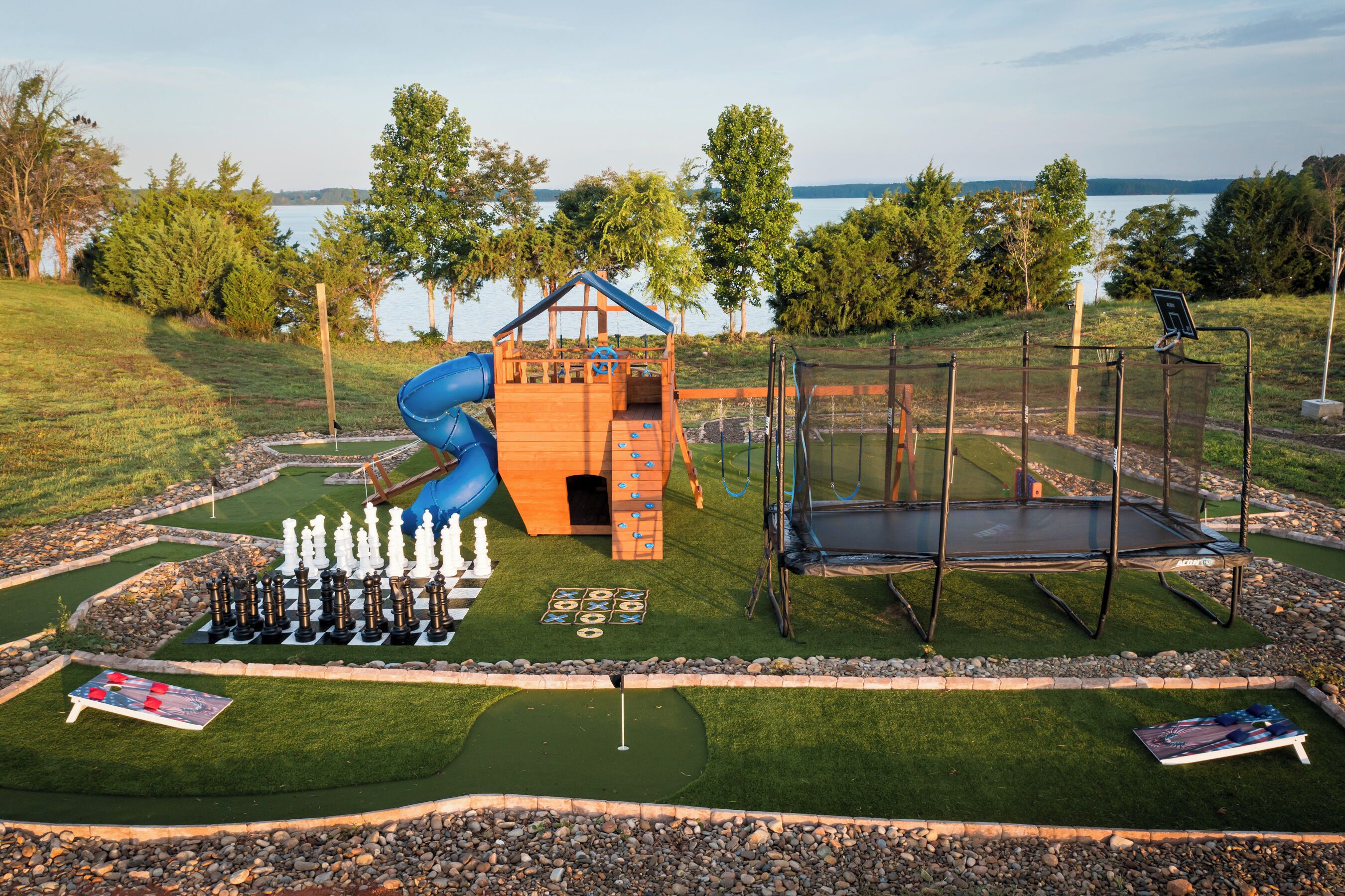 A playground with a slide, climbing wall, and trampoline, surrounded by a golf course and a lake view.