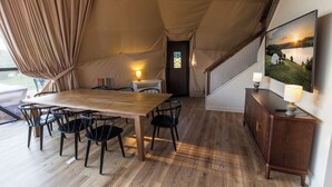 Alojamento em tenda com mesa de jantar de madeira, cadeiras, um pequeno armário branco e uma pintura de paisagem.