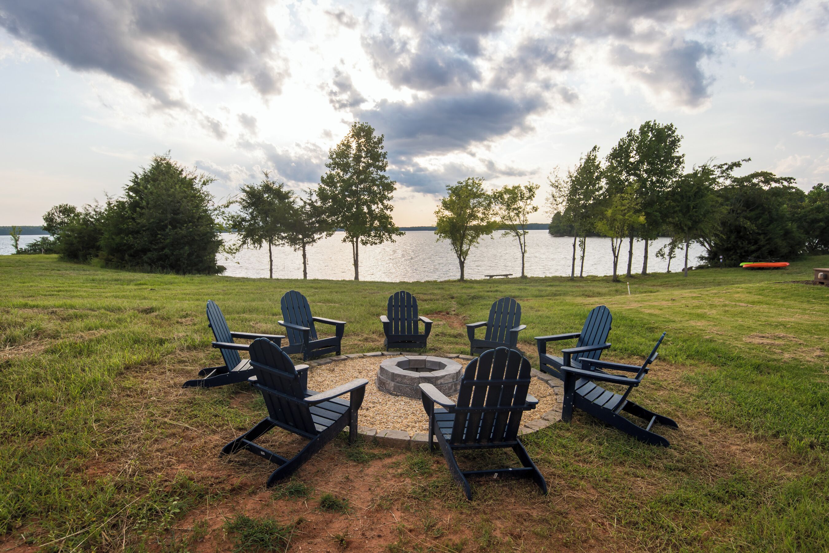 Un espace extérieur aménagé au bord du lac, avec des chaises Adirondack disposées autour d’un foyer circulaire.