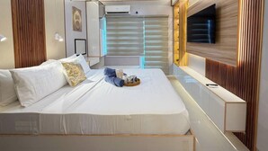 A modern bedroom with a large bed, bedside tables, a TV mounted on the wall, and a window with blinds. - Room-style Studio Unit in Smdc (Iloilo)