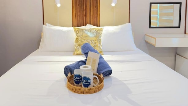 A neatly made bed with white linens, a blue towel, a wicker tray with a mug and a bottle, and a pillow with a leaf pattern. - Room-style Studio Unit in Smdc (Iloilo)
