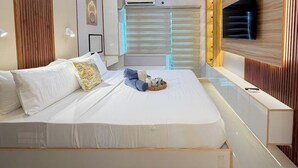 A modern hotel room with a large bed, a bedside table, a built-in TV, and a window with blinds. - Room-style Studio Unit in Smdc (Iloilo)