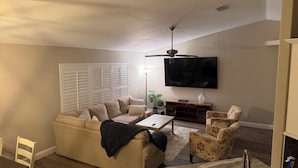 Una sala de estar con un sofá color beige, un sillón estampado, una mesa de centro, una lámpara de pie y un televisor de pantalla plana montado en la pared.