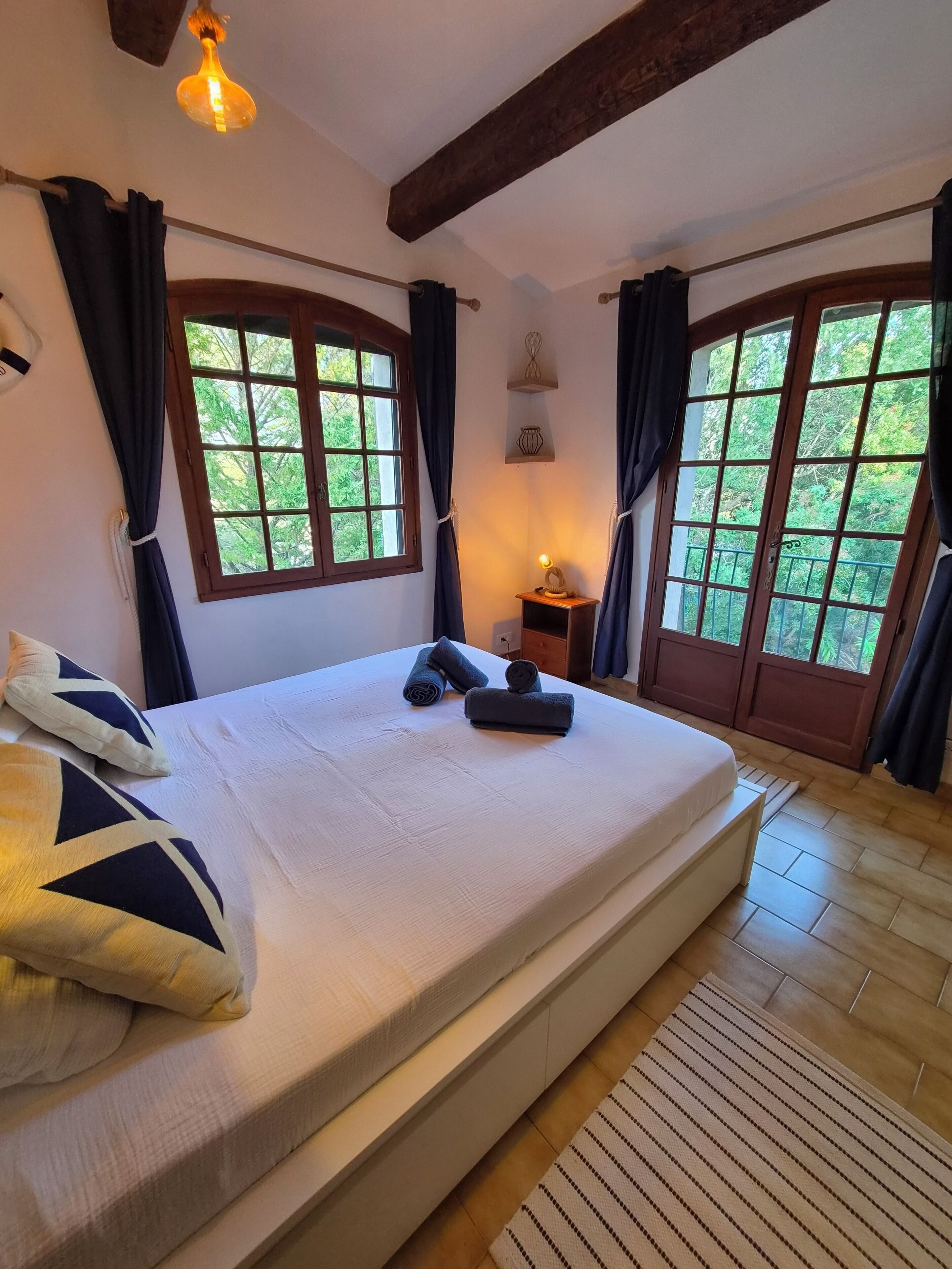 Een slaapkamer met een groot bed, twee ramen met gordijnen, een bedlampje en een klein tafeltje met een lamp.