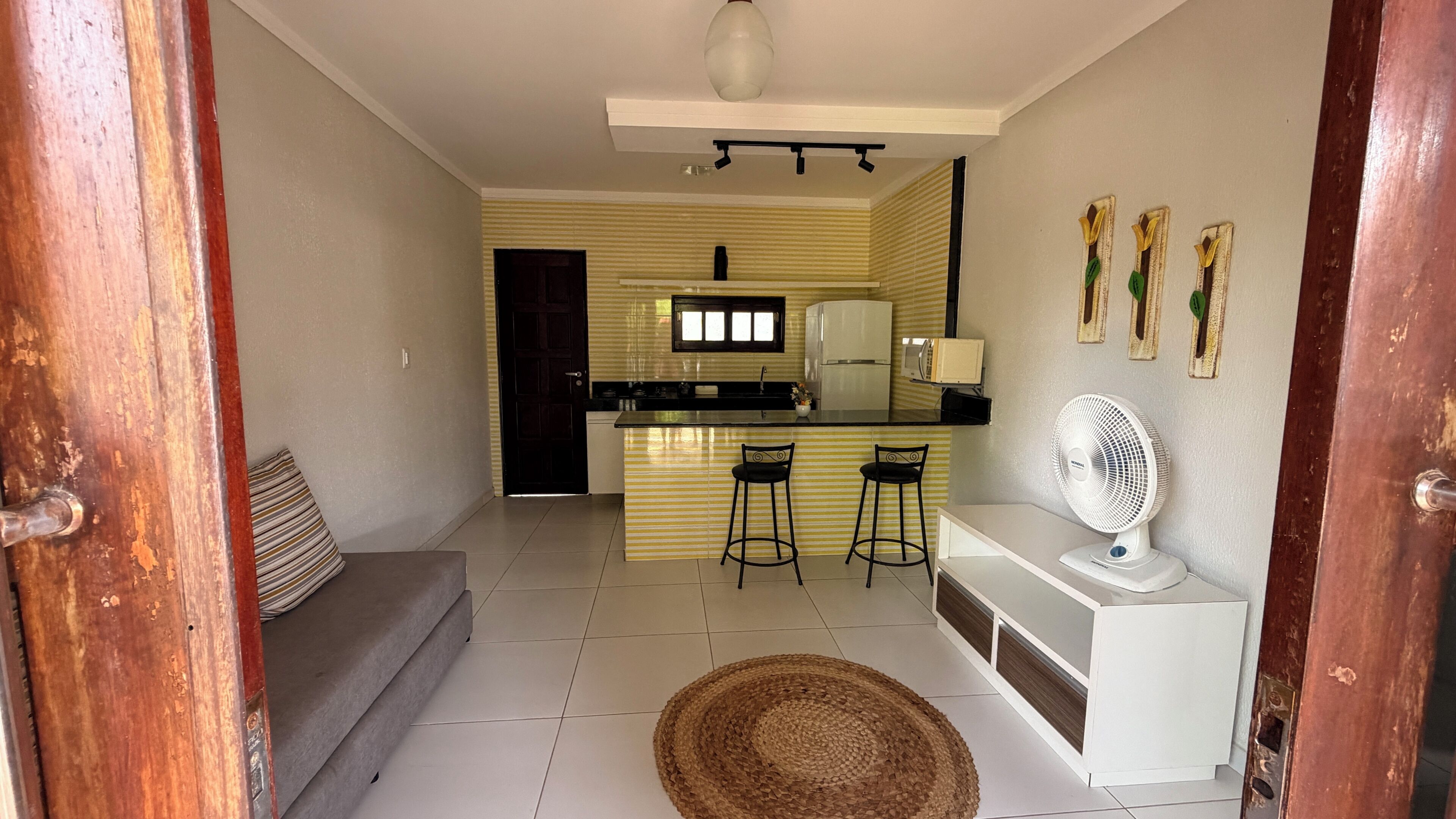 Un espacio habitable funcional con kitchenette, un sofá, dos taburetes tipo bar, un ventilador de pie y un tapiz tejido.