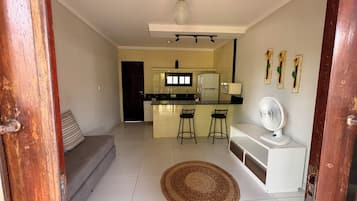 Un espacio habitable funcional con kitchenette, un sofá, dos taburetes tipo bar, un ventilador de pie y un tapiz tejido.