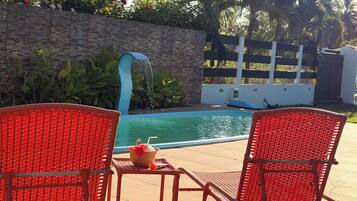 Área ao redor da piscina com duas cadeiras de vime vermelhas, uma mesa pequena com bebida e cesta, e uma parede de pedra com plantas.