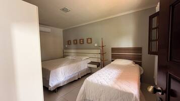 Quarto com duas camas de solteiro, cabeceira de madeira, ar condicionado fixado na parede e porta com painel de vidro.
