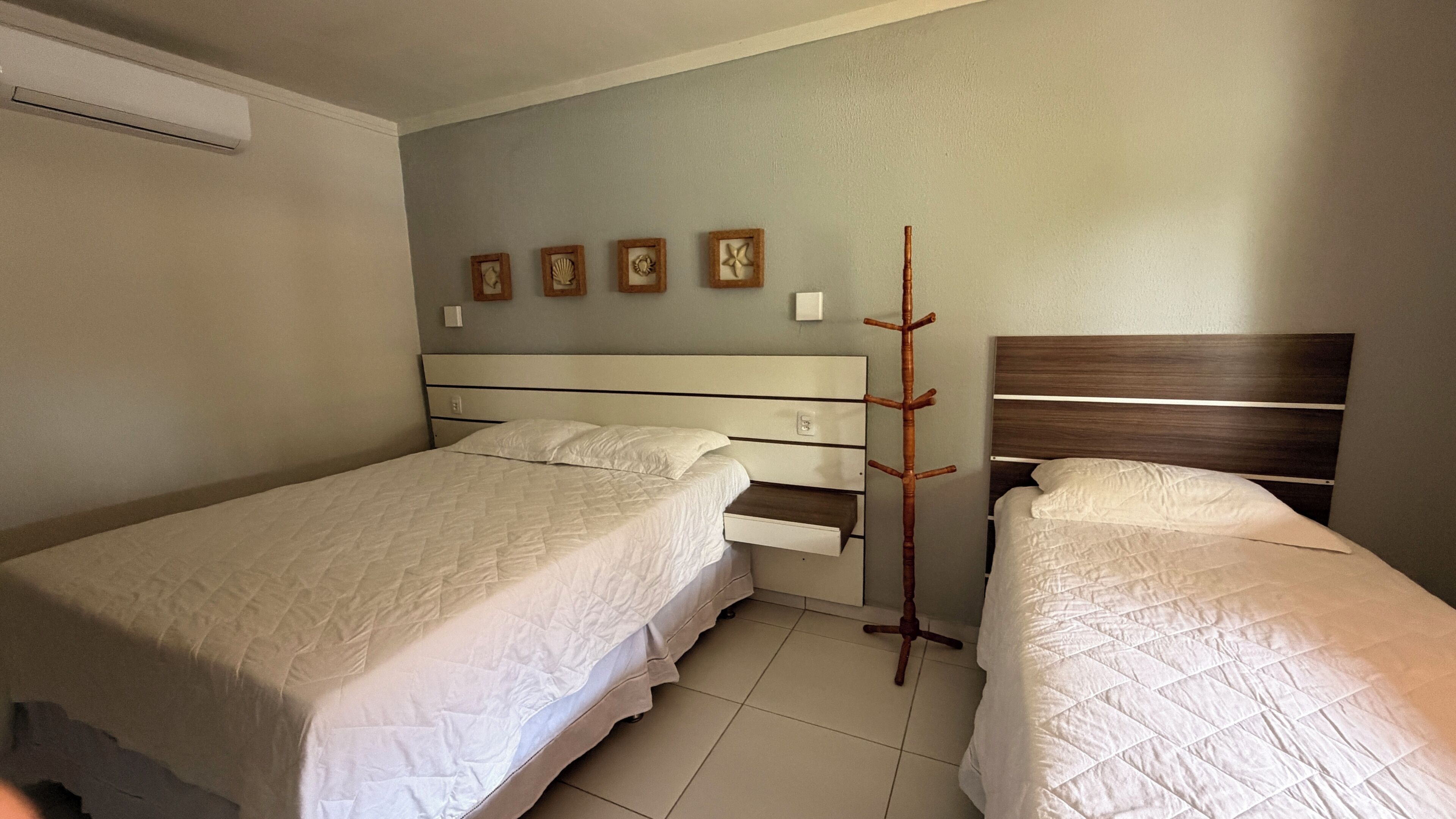 Ein Hotelzimmer mit zwei Betten, einem hölzernen Kopfteil, einer Wand-Klimaanlage und einer Wand mit vier gerahmten Bildern.