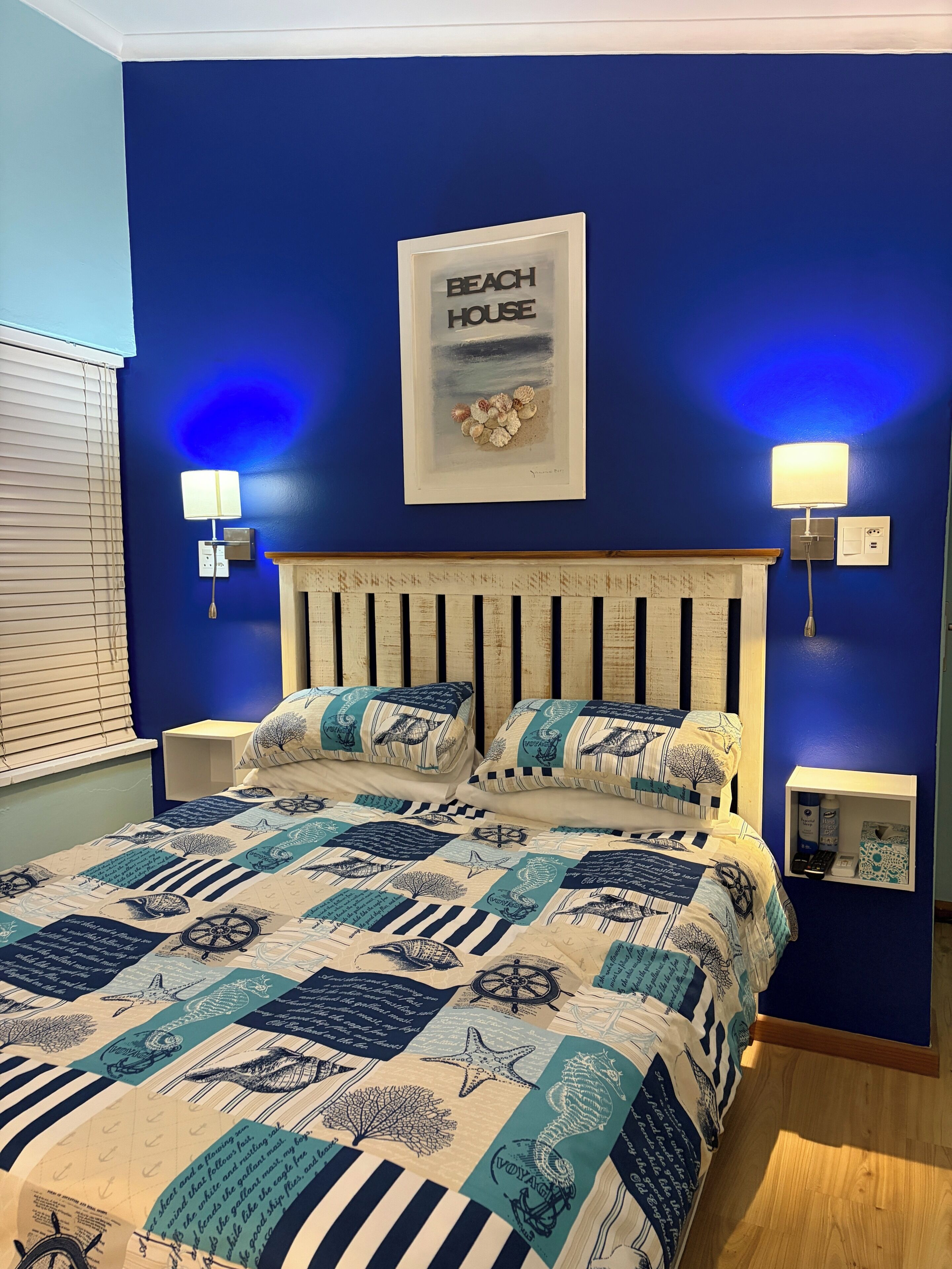A bedroom with a blue wall, a bed with nautical-themed bedding, a framed picture of a beach house, and wall-mounted lamps.